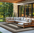 A modern outdoor sectional sofa with light beige cushions and a matching ottoman, arranged on a large patterned area rug on a covered patio. The setting includes a stone wall, black-framed glass doors, and a lush green garden in the background.