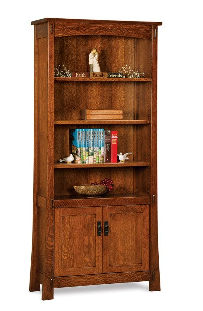 A tall wooden bookcase with three open shelves and two cabinet doors below, shown against a plain white background. The bookcase is made of medium brown wood with visible grain, and is styled with books, small figurines, and a bowl.