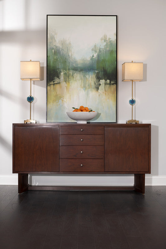 Wooden sideboard with a bowl of oranges, two lamps, and a large abstract painting on a white wall.