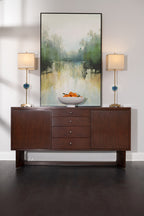 Wooden sideboard with a bowl of oranges, two lamps, and a large abstract painting on a white wall.
