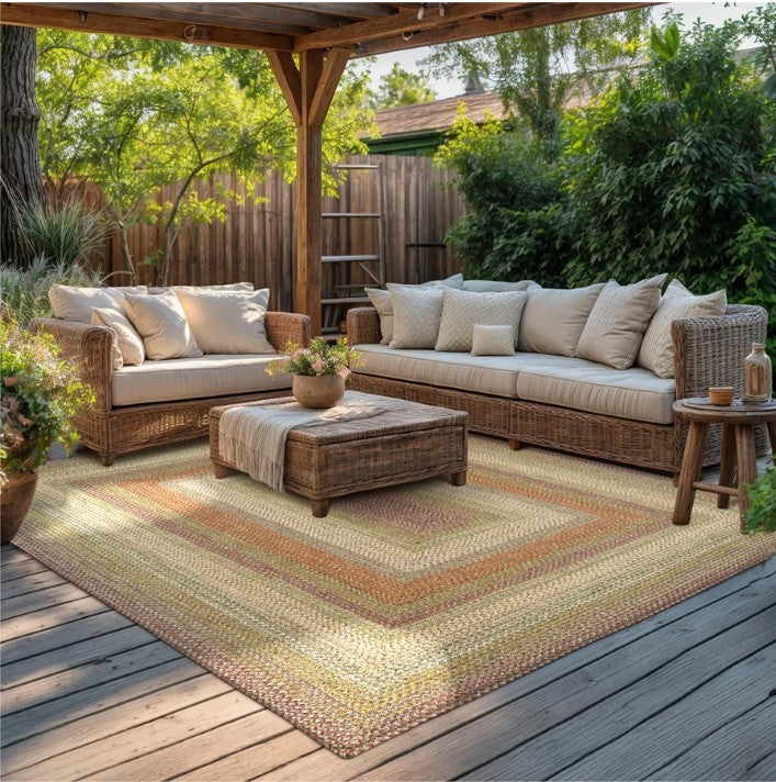 Outdoor patio furniture set on a wooden deck under a pergola, featuring a wicker sofa, matching loveseat, woven ottoman, and a large braided area rug in warm neutral tones.