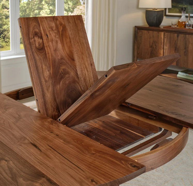 A close-up of a wooden dining table with a lift-top extension mechanism open, showing the rich brown wood grain in a bright dining room setting.