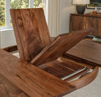 A close-up of a wooden dining table with a lift-top extension mechanism open, showing the rich brown wood grain in a bright dining room setting.