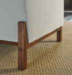 Close-up of a light beige upholstered chair or sofa leg with a dark brown wooden frame and textured woven rug underneath.