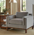 A light gray upholstered armchair with a dark wood frame, shown in a styled living room setting with a wooden side table, lamp, and windows.