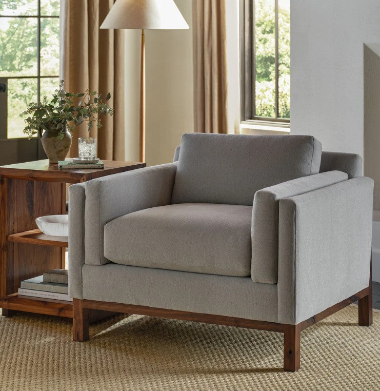 A light gray upholstered armchair with a dark wood frame, shown in a styled living room setting with a wooden side table, lamp, and windows.