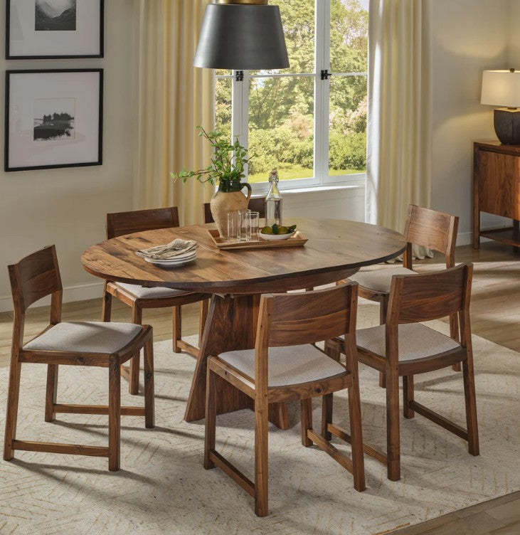 A round wooden dining table with six matching wooden bar-height chairs featuring light beige upholstered seats, set in a bright, modern dining room.