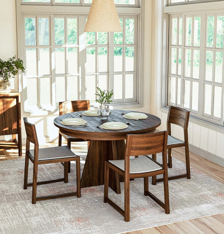 A round wooden dining table with four matching wooden chairs in a bright, sunlit dining room with large white-framed windows.