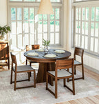 A round wooden dining table with four matching wooden chairs in a bright, sunlit dining room with large white-framed windows.