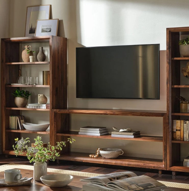A dark brown wooden media console and shelving unit set, styled with books, vases, and a television mounted on the back wall in a warm, sunlit room.