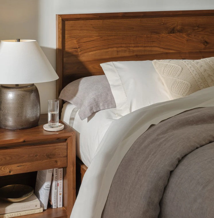 A close-up of a bed with white and gray bedding, a wooden headboard, and a matching wooden bedside table holding a lamp, a glass of water, and books.