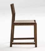 A modern wooden barstool with a dark brown wood frame, a light beige woven seat cushion, and a tall, minimalist backrest, shown from a side profile view on a light gray background.