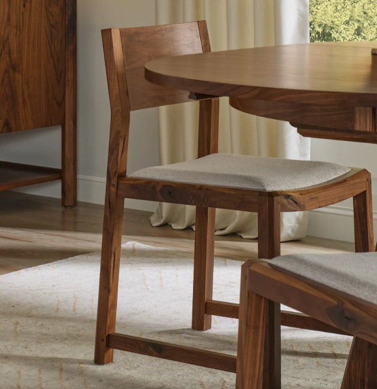 A wooden dining chair with a light beige upholstered seat cushion, shown in a real indoor room beside a wooden table and rug.