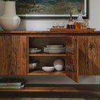 A warm medium-brown wooden sideboard cabinet with one door open, showing stacked white plates and bowls inside. The cabinet is styled in a home setting with a white vase, glasses, and a decanter on top, and a framed landscape print on the wall behind it.