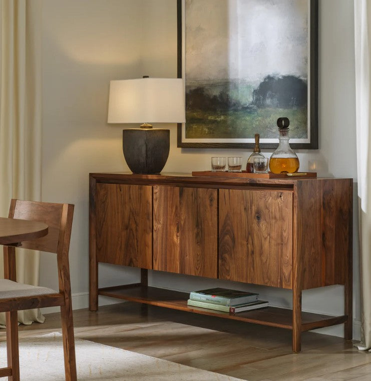 A mid-century modern wooden sideboard or credenza in a warm medium brown finish, styled in a dining room setting with a table lamp, glasses, a decanter, books, and framed wall art.