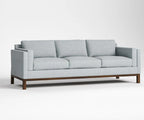 A three-seat sofa with light gray fabric upholstery and a dark brown wooden base, shown against a plain light gray studio background.