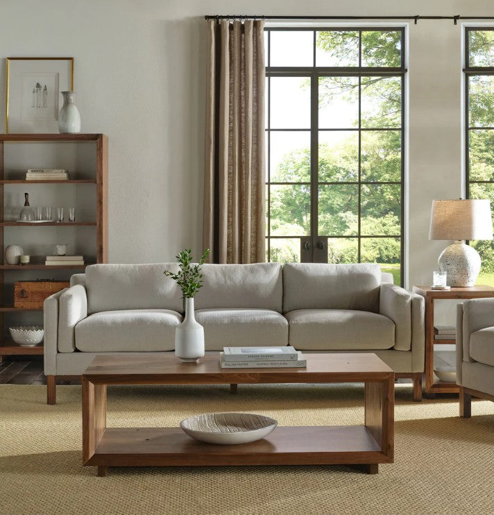 A light beige upholstered three-seat sofa in a styled living room, positioned in front of a wooden coffee table and a dark wood shelving unit. The room features large black-framed windows with green foliage outside, beige curtains, and a textured beige rug.