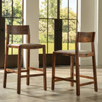 Two wooden bar stools with light beige upholstered seats, shown in a bright interior with large windows and a view of greenery outside.