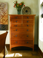 A six-drawer wooden chest of drawers with a warm reddish-brown finish and dark round knobs, styled in a room with a vase of flowers and a decorative plate on top.