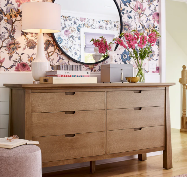 A light oak wood six-drawer dresser in a beautifully styled bedroom, decorated with a white table lamp, books, a gray box, and a vase of pink flowers against a floral wallpaper background.