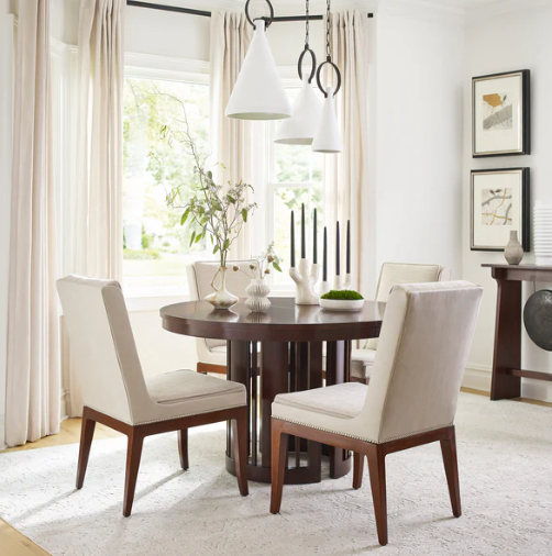 A round dark wood dining table surrounded by four upholstered dining chairs in a bright, modern dining room. The chairs have cream-colored fabric seats and backs with dark wood legs.