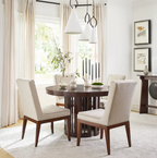 A round dark wood dining table surrounded by four upholstered dining chairs in a bright, modern dining room. The chairs have cream-colored fabric seats and backs with dark wood legs.