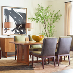 A mid-century modern dining room featuring a rectangular wooden table with a slatted pedestal base, surrounded by high-back upholstered chairs in dark gray and sage green. The room has beige walls, a large black and white abstract painting, two table lamps, and a tall green plant.