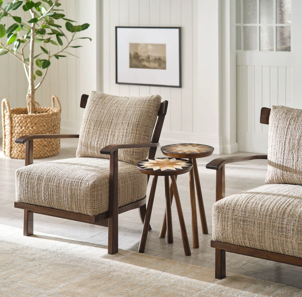 A styled interior scene showing two upholstered armchairs with dark wood frames and a small round side table between them, set in a bright room with a plant, framed wall art, and paneled wall.