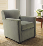 A single upholstered armchair in a muted sage green fabric, shown in a bright living room setting with wood flooring, a woven rug, a side table, and a vase of white flowers.