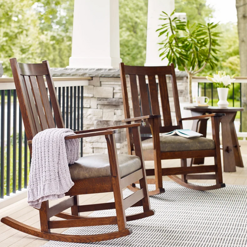 Two dark brown wooden rocking chairs with cushioned seats on a porch, with a light purple knit throw blanket draped over the left chair. A small wooden side table sits between them, and a stone wall and green trees are visible in the background.