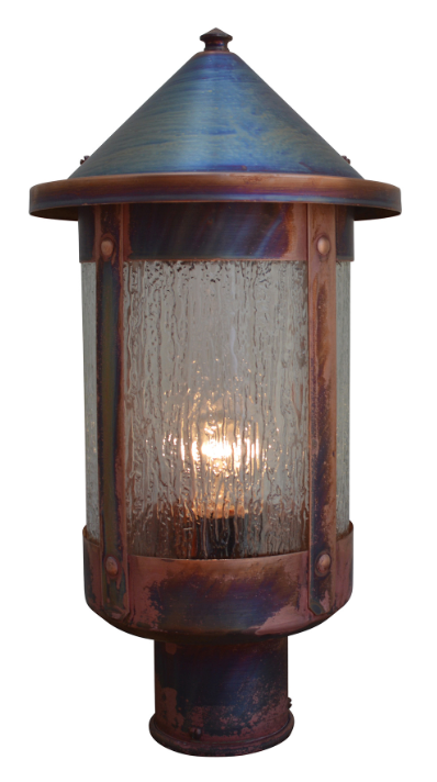 A rustic outdoor post cap light fixture with a distressed copper and blue-purple patina finish, featuring a textured clear glass shade and a warm glowing bulb inside, isolated on a white background.