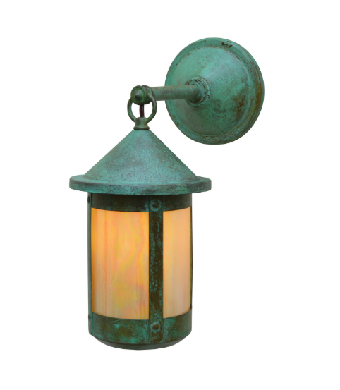 A single outdoor wall sconce with a distressed green patina finish, featuring a cylindrical glass shade that glows with a warm orange light, mounted on a round wall plate against a white background.