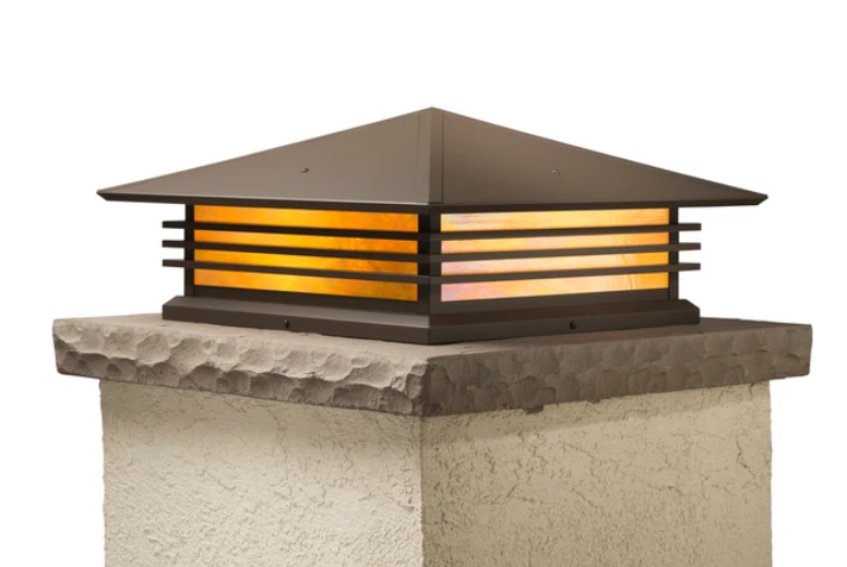 A dark bronze square outdoor light fixture with horizontal slats and a warm glowing amber glass panel, mounted on a textured stone pillar against a white background.