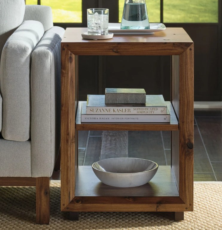 A rustic wooden end table with glass-front shelves, styled with books, a bowl, and a drink, positioned next to a light gray armchair in a home interior.